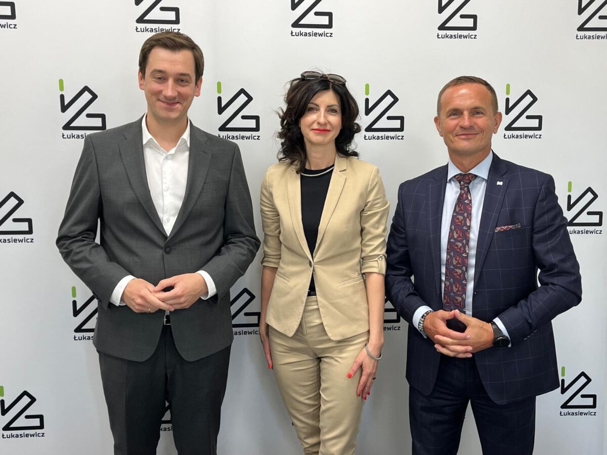 Mężczyzna, kobieta, mężczyzna na tle ścianki z logotypem Łukasiewicza