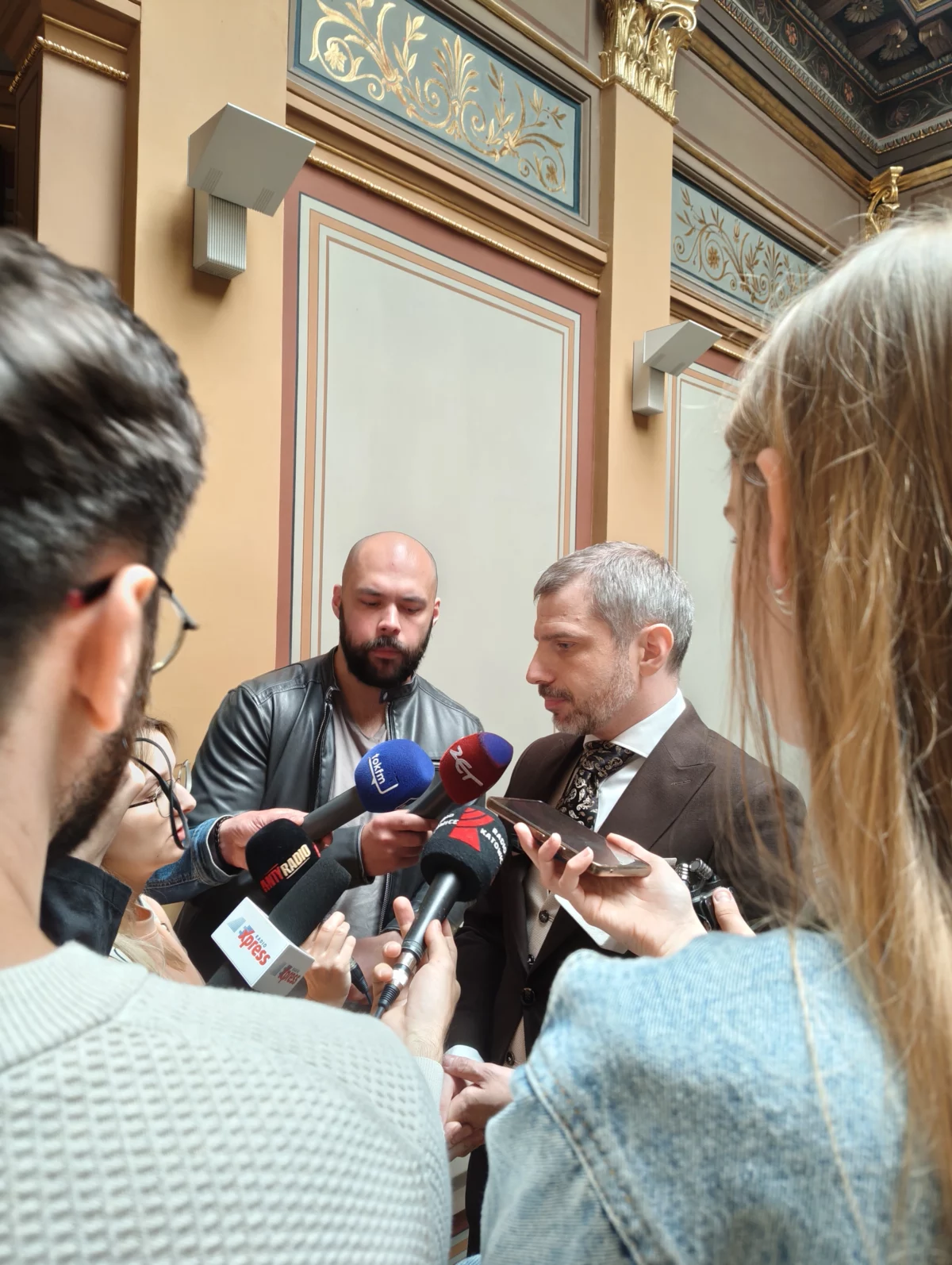 Mężczyzna w garniturze w otoczeniu dziennikarzy z mikrofonami