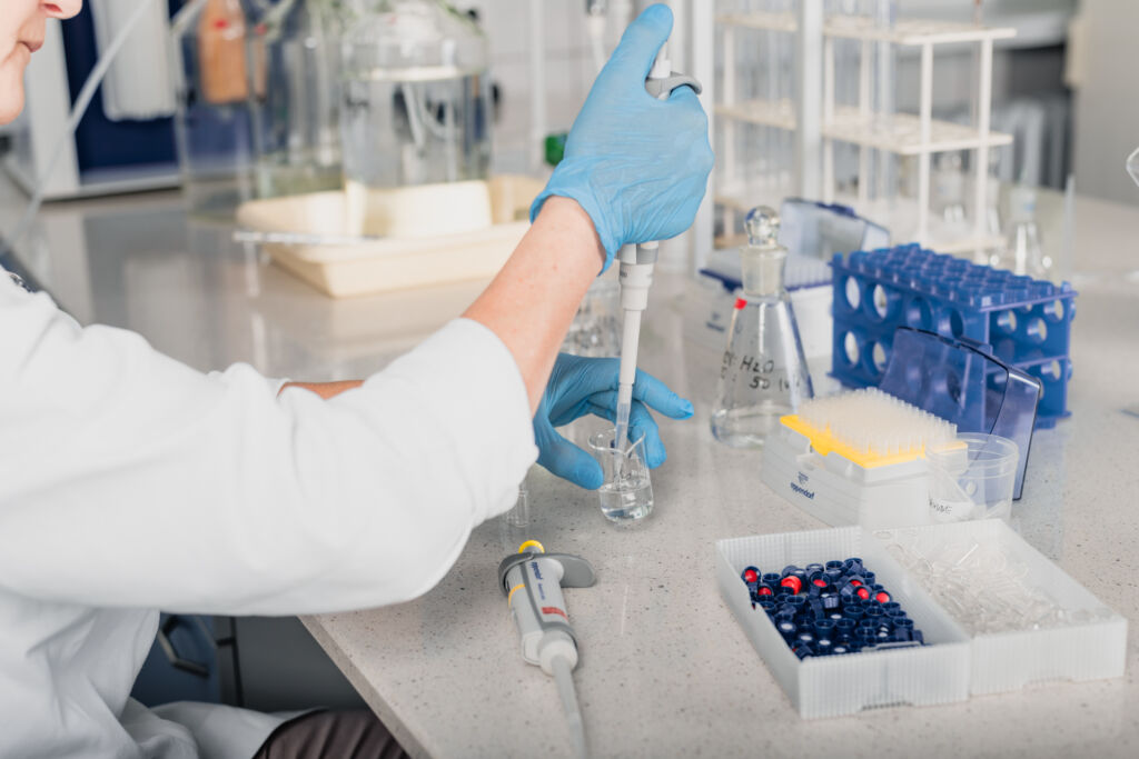 Na zdjęciu widać prace w laboratorium. Dłonie ubrane w rękawiczki lateksowe obsługują automatyczną pipetę.